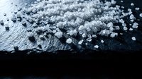 Close-up of white crystalline salt granules scattered on dark surface, suitable for food, seasoning, mineral, and culinary content.