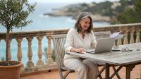 Mature blonde businesswoman working on laptop at Mediterranean coastal terrace with sea view, remote work lifestyle concept for professionals.
