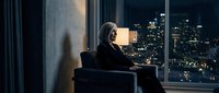 Mature businesswoman sitting by office window overlooking city skyline at night, contemplating urban landscape with illuminated buildings in background.