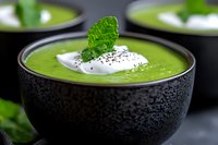 Green pea soup garnished with sour cream, chia seeds and fresh mint leaf served in black bowl. Condensation droplets add appetizing appeal.