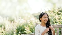 Young woman with long dark hair and South Asian ethnicity, wearing casual beige clothing, holding microphone outdoors in garden setting with blooming white flowers and green foliage, suitable for event coverage, speaking engagements, or outdoor performance concepts.