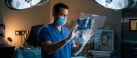 Male surgeon in blue scrubs and medical mask examining X-ray film in modern operating room with surgical lights overhead for medical diagnosis.