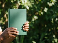 Person holding green hardcover book outdoors with blurred foliage background, suitable for education, reading, literature, and publishing concepts.