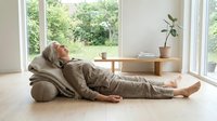 Senior woman with gray hair, Caucasian ethnicity, relaxing on floor with cushions in bright, minimalist living room with large windows and wooden furniture.