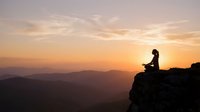 Woman meditating on mountain cliff edge during golden sunset with layered hills in background creating peaceful mindfulness scene for wellness content.
