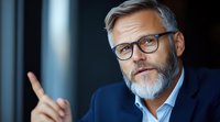 Confident mature businessman with gray beard and glasses gesturing while speaking, wearing navy suit and white shirt against dark background. Professional corporate portrait.