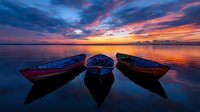 Wooden fishing boats moored at sunset with dramatic orange and purple sky reflecting in calm water, creating mirror effect and peaceful atmosphere at dusk.