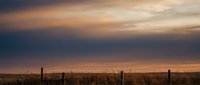 Rural countryside landscape with wooden fence posts and golden grass field under dramatic sunset sky with orange and purple clouds creating peaceful agricultural scenery.