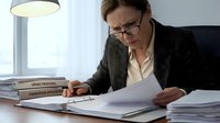 Middle-aged Caucasian businessman in glasses and suit reviewing legal documents at office desk with stacks of papers and modern lamp in background.