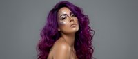 Young woman with vibrant purple curly hair and glamorous makeup posing confidently against gray background for beauty and fashion concepts.