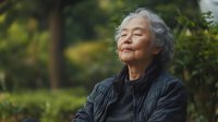 Senior Asian woman with gray hair sitting peacefully outdoors with eyes closed and gentle smile, wearing black quilted jacket against natural green garden background.