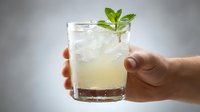 Refreshing mint cocktail with ice in glass held by hand against gray background. Cold summer drink with fresh green mint leaves for bar menu design.