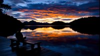 Silhouette of person sitting on wooden pier at sunset, dramatic orange and blue sky reflecting in calm lake water surrounded by mountain landscape.