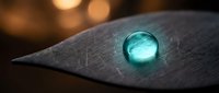 Glowing turquoise water droplet on dark textured leaf surface with warm bokeh lights in background creating magical nature scene for wellness designs.