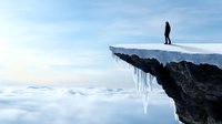 Lone silhouetted figure stands at edge of snow-covered cliff with dramatic icicles hanging above misty cloud-filled valley, expressing solitude and adventure.