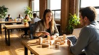 Young professional woman and man having business meeting in modern coworking office space with natural lighting and plants in background.