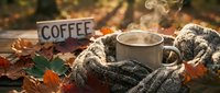 Steaming mug wrapped in cozy knit scarf surrounded by colorful autumn leaves on rustic wooden table outdoors, warm fall coffee moment.