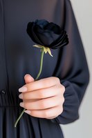 Female hand holding single black rose against dark background, wearing black clothing. Dramatic gothic romantic mood for luxury design and art projects.