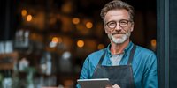 Mature male small business owner with tablet standing at cafe entrance, bokeh lights in background.