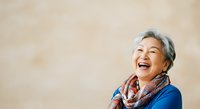Senior Asian woman with gray hair wearing blue top and colorful scarf, smiling joyfully against neutral beige background for lifestyle and wellness content.