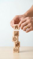 Hands protecting wooden blocks with financial icons representing investment growth, real estate planning, and wealth management strategy on neutral background.