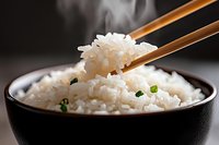 Steaming white rice held by wooden chopsticks above a black bowl filled with cooked rice and green onion garnish for Asian cuisine cooking.