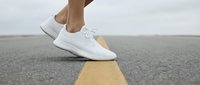 Person wearing white sneakers standing on yellow road line marking on asphalt pavement, representing journey, direction, and forward movement for lifestyle and travel concepts.