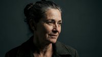 Middle-aged woman with dark hair styled in a bun, appearing to be of Caucasian ethnicity, wearing a dark olive or charcoal jacket against a neutral gray background. Her expression is contemplative and serene, suitable for corporate, editorial, or lifestyle content.