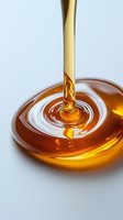 Golden honey or syrup stream pouring and creating concentric ripples on glossy amber liquid surface against light background. Macro food photography.
