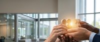 Diverse team of professionals joining hands around glowing light bulb in modern office, symbolizing innovation, collaboration, and shared ideas for business success and teamwork.