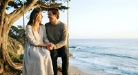 Young couple embracing on beach at sunset under tree, romantic moment by ocean waves, love and connection in natural coastal setting for lifestyle content.