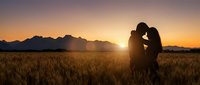 Romantic couple silhouette embracing in golden wheat field at sunset with mountain backdrop creating warm intimate atmosphere for love concepts.