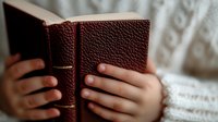 Hands holding open brown leather book with textured cover against soft white background, capturing intimate reading moment in natural lighting.