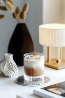 Iced latte with cream in textured glass on striped coaster, styled with modern minimalist decor including black vase, white ceramic, and table lamp on white surface.