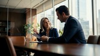 Middle-aged Caucasian woman and man in business attire discussing work matters at conference table in modern office with city skyline view through windows.