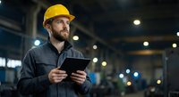 Industrial worker with yellow hard hat using digital tablet in manufacturing facility with blurred lights background for safety and technology concepts.