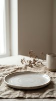 Minimalist ceramic plate on natural linen fabric with dried botanicals and cup, neutral beige tones, soft window light, rustic table setting for home decor and lifestyle content.