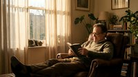 Middle-aged Caucasian man with glasses reading book in cozy armchair by sunny window. Warm indoor lighting creates peaceful home library atmosphere.