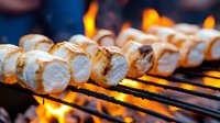 Roasted marshmallows on metal skewers over glowing campfire flames, toasted to golden brown with melting edges against dark background for outdoor cooking.