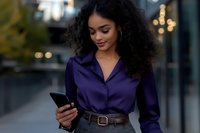 Young African American businesswoman with natural curly hair checking mobile phone while walking outdoors in urban setting, wearing navy blue shirt and leather belt.