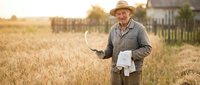 Senior Caucasian male farmer in straw hat holding tablet and documents in golden wheat field at sunset for agricultural technology and farming business.