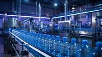 Automated conveyor line with blue plastic bottles moving through modern beverage manufacturing facility with industrial machinery and blue lighting.