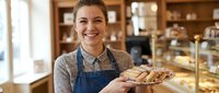 Young female baker with medium complexion and brown hair, wearing blue apron and beige cardigan, smiling while holding fresh baked pastries in bakery shop with wooden shelves and display cases.