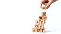 Hand placing wooden cube with growth chart icon on stack of business symbol blocks representing strategic planning and success building.