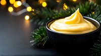 Creamy yellow custard or lemon curd in black bowl with Christmas fir branches and golden bokeh lights creating festive holiday dessert scene for seasonal baking.