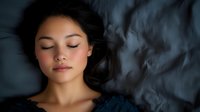 Young Asian woman sleeping peacefully on dark silk bedding, face illuminated by soft moody lighting, expressing serenity and wellness for health and lifestyle concepts.