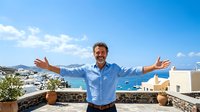 Man in blue shirt with arms outstretched on sunny terrace overlooking coastal landscape with mountains, expressing joy and freedom during vacation or travel.