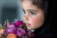 Young girl in black headscarf holds colorful bouquet with roses and carnations, side profile portrait showing innocent expression and natural makeup against dark background.
