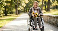Smiling mature Caucasian woman in wheelchair holding colorful autumn flowers in sunny park setting. Disability awareness, accessibility, senior lifestyle concept.