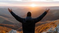 Man with raised arms celebrating freedom and success on mountain peak during golden hour sunset with panoramic valley view below.